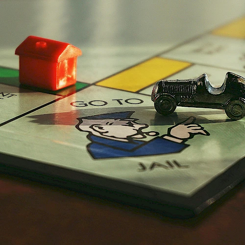 A red toy house and a small toy car sit on a Monopoly board near a jail space, with a cartoon figure peeking up from the jail.