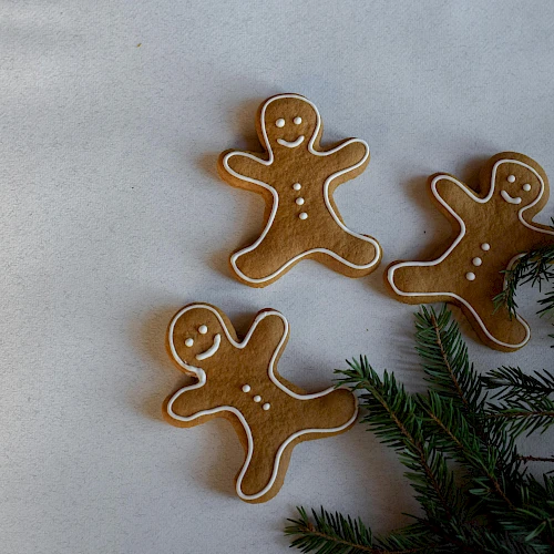 Three gingerbread cookies shaped like little people, with white icing faces and buttons, placed on a light surface beside green pine branches.