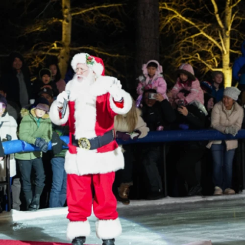 A man dressed as Santa Claus stands on an ice rink, with a crowd of people watching behind a barricade at night.