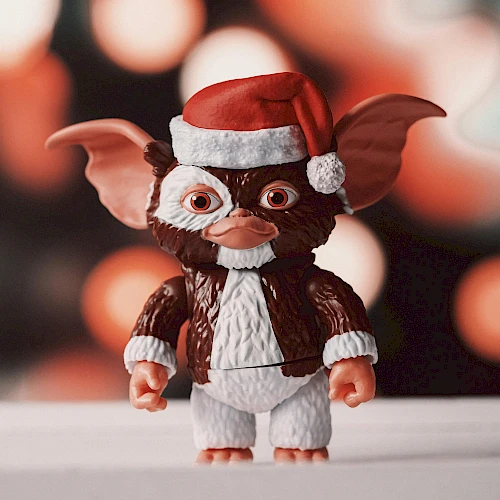 A tiny Gizmo-like creature wearing a Santa hat and white fur-trimmed suit stands on a surface, with warm blurred red bokeh lights behind.